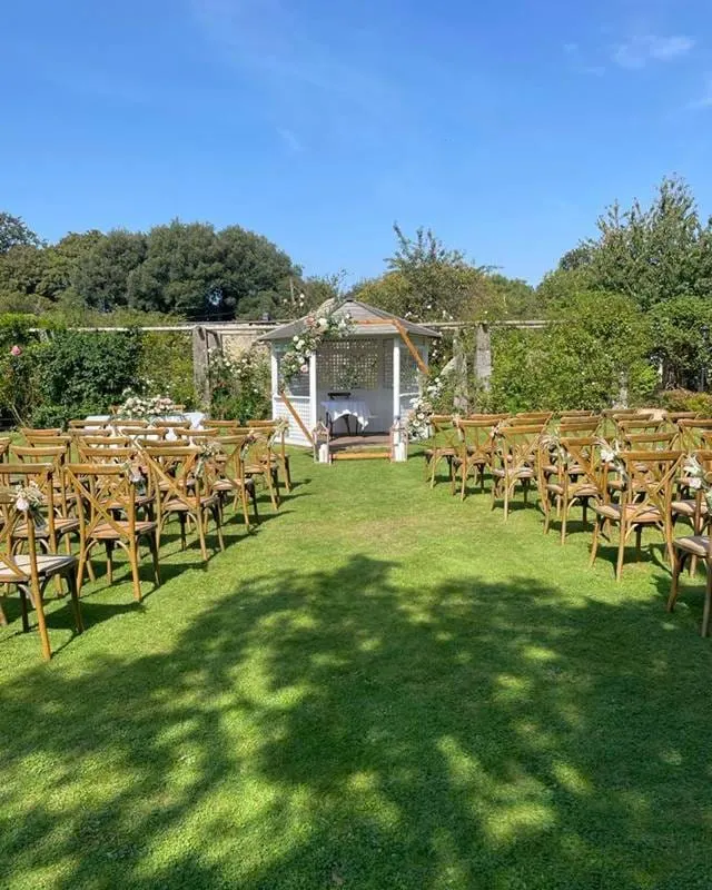 Outdoor garden ceremony setup with wooden chairs arranged on a lawn facing a decorated arbour, surrounded by trees.