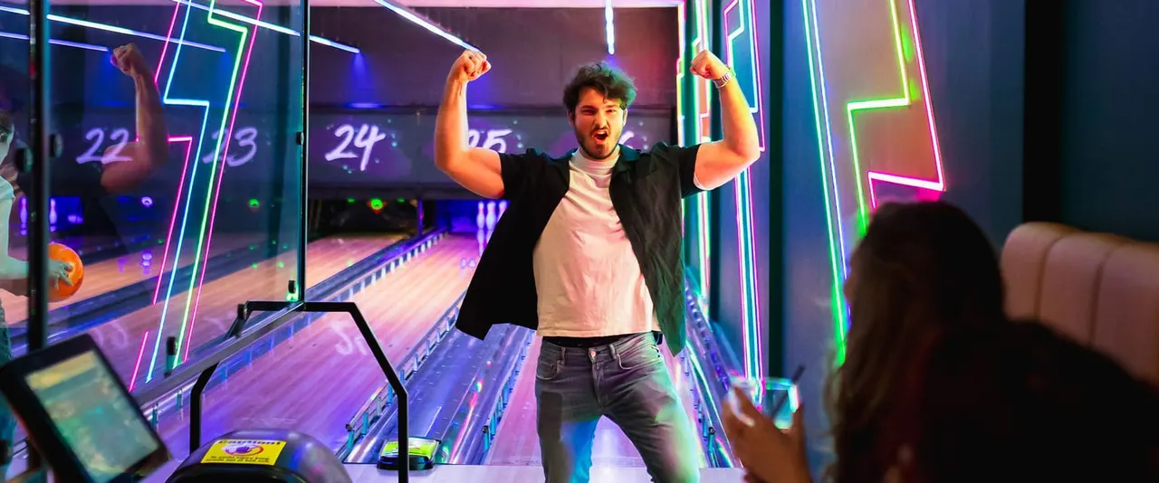 Man celebrating a bowling strike with arms raised at a neon-lit bowling lane.