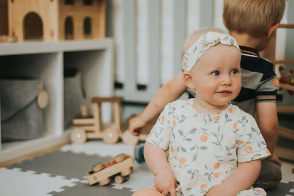 Two young children play together in an indoor play space surrounded by wooden toys and storage units.