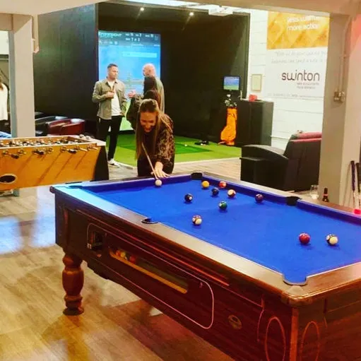 A woman plays pool at a leisure venue with a golf simulator and other activities in the background.