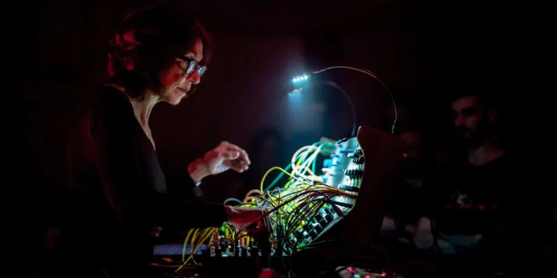 Person wearing glasses operating electronic synthesiser equipment with illuminated cables in darkened venue.
