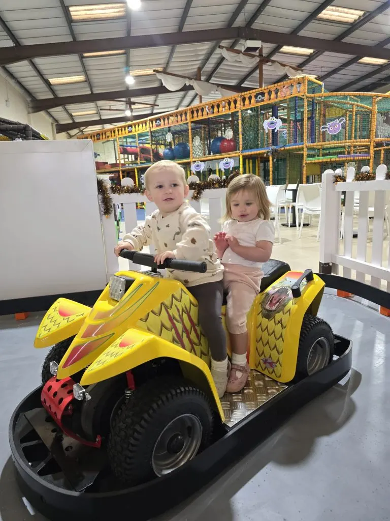 Two toddlers ride together in a yellow dragon-shaped bumper car at an indoor play centre.