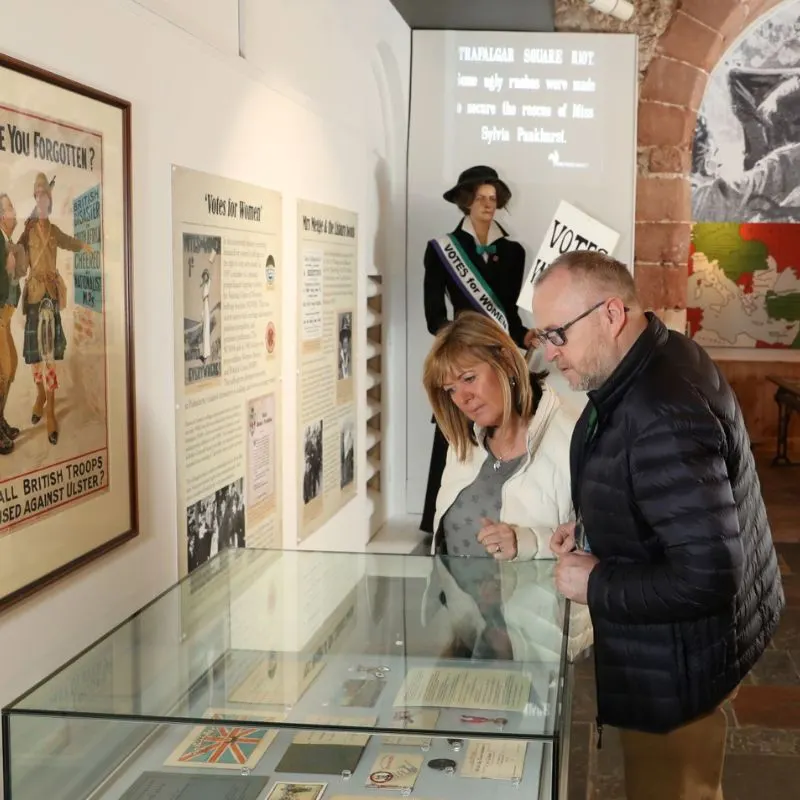 Two visitors examine a glass display case containing historical documents and artefacts in a museum exhibition about Irish history and linen.