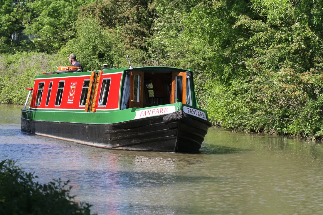 A red and green narrowboat moored on a tree-lined canal with a person visible on the roof.