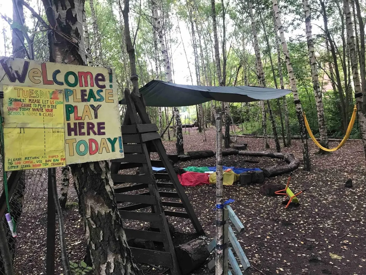 Woodland outdoor play area with welcome sign, climbing ladder, rope swing and scattered play equipment amongst birch trees.