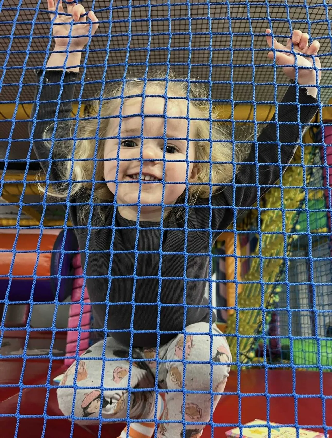 Child playing in an indoor soft play facility with climbing nets and coloured panels.