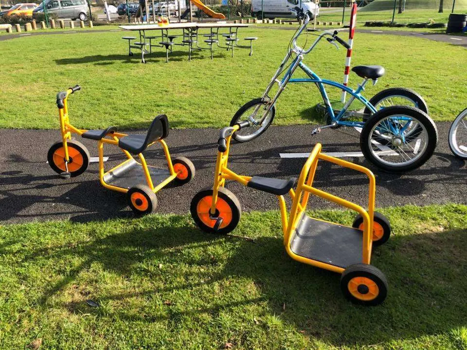 Colourful children's tricycles and a blue trike parked on a path beside a grassy play area with picnic tables.