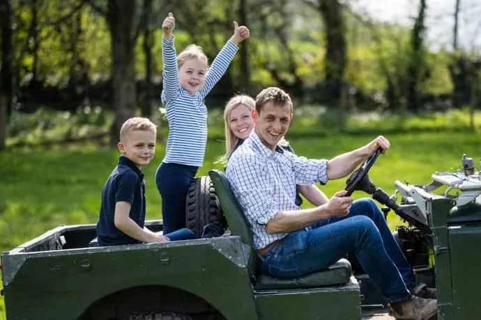 Family of five riding in a trailer towed behind a farm vehicle through green parkland.
