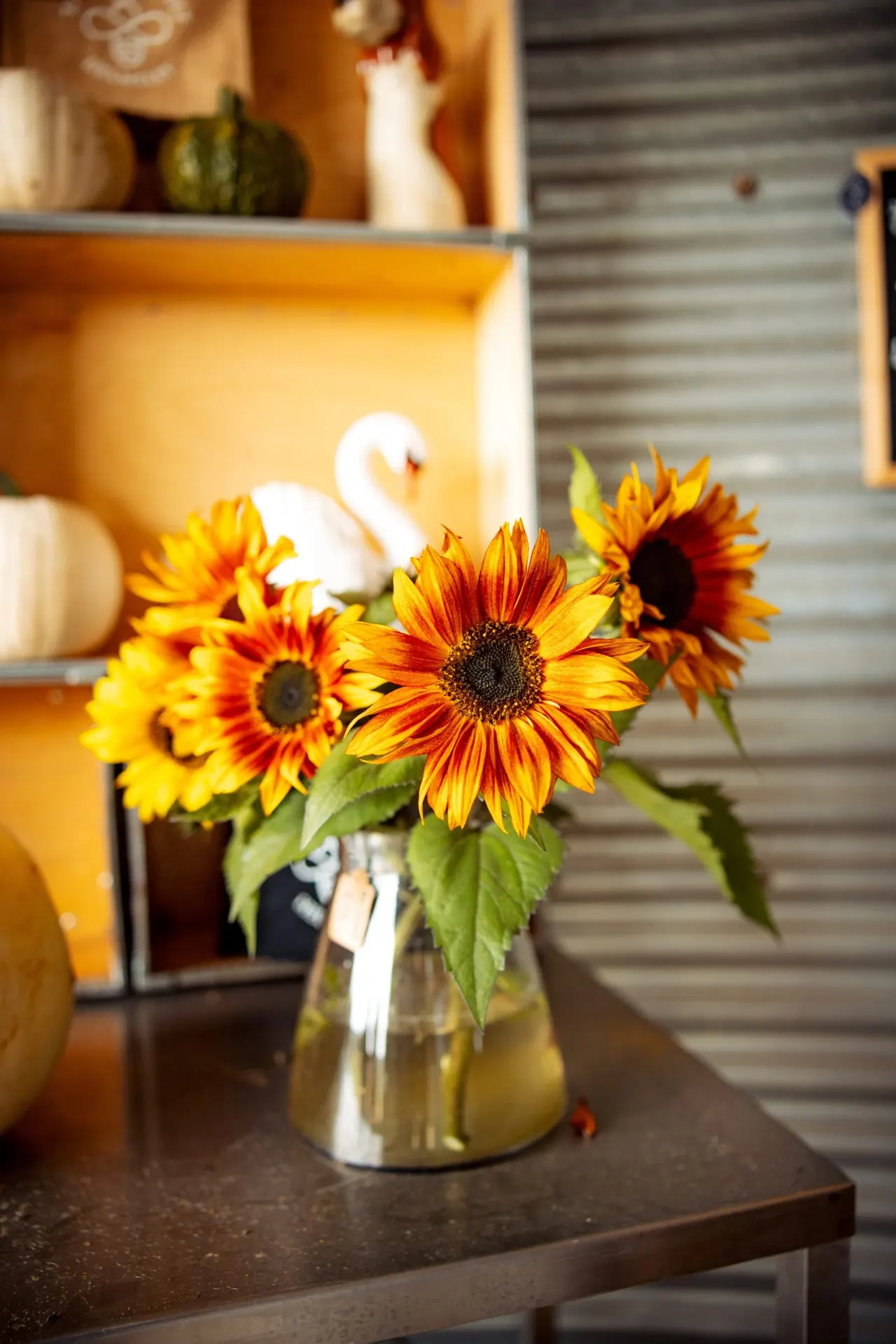 Red and yellow sunflowers in a glass vase on a dark table, with yellow shelving and decorative items in the background.