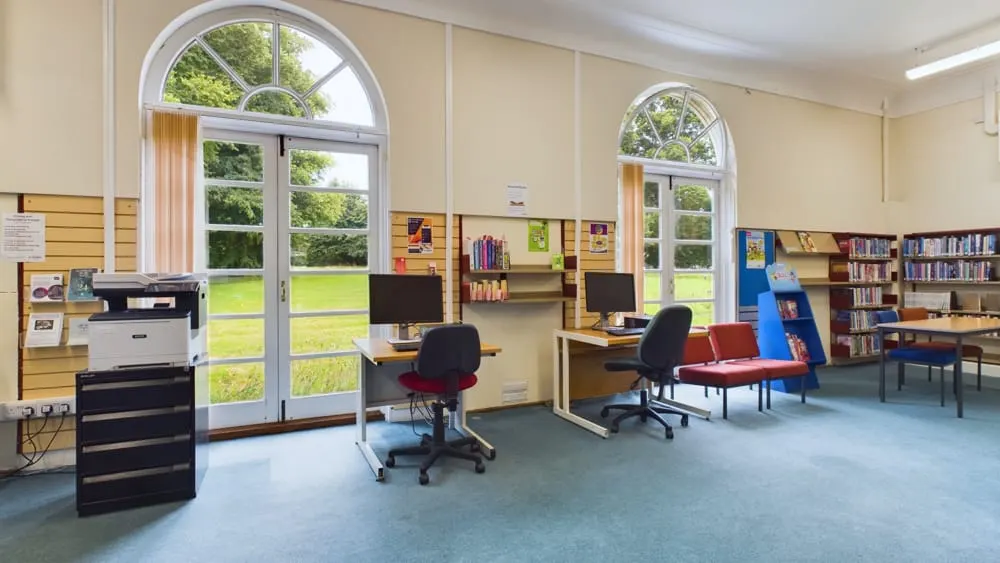 Interior of a bright library space with computer workstations, bookshelves, seating areas and views through large arched windows to green grounds outside.