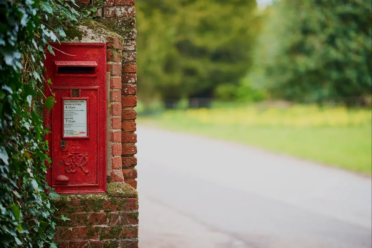 Red postbox mounted on a brick wall covered with ivy, beside a tree-lined driveway.