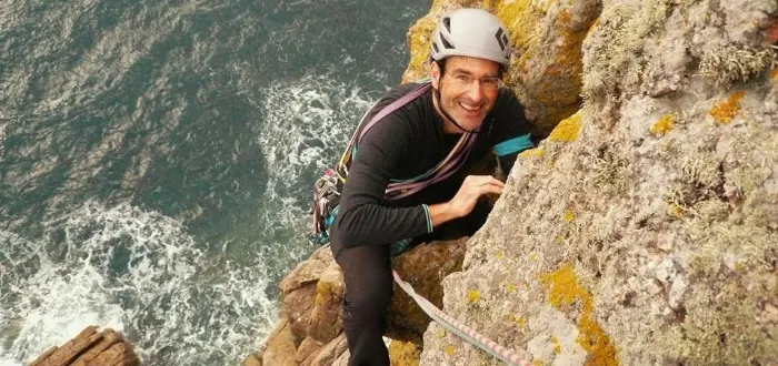 Man rock climbing on a seaside cliff face with safety helmet and rope, smiling at camera above turbulent sea.