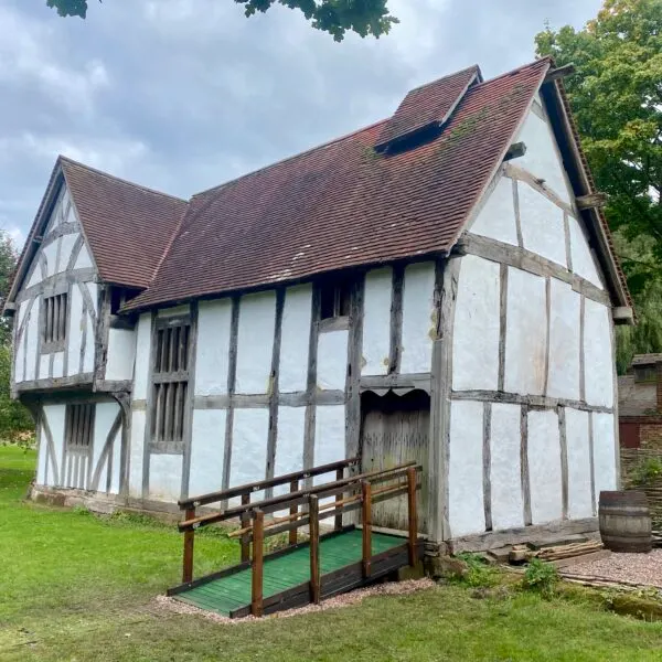 Historic timber-framed white and black building with steep tiled roofs and wooden entrance ramp set in grassy grounds.