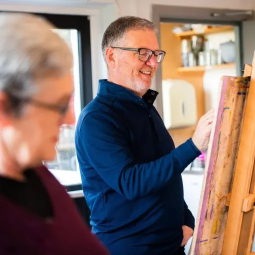 Man in blue shirt and glasses painting on an easel whilst another person watches.