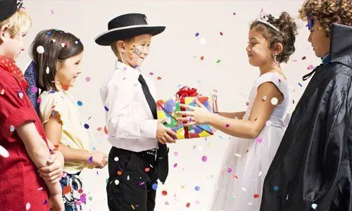 Children in costume at a party exchange a colorful gift whilst confetti falls around them.