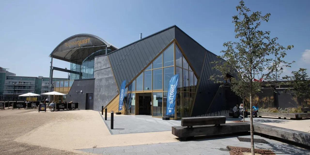 Modern building with large windows and gold framing at a waterfront science centre, with sandy plaza and seating areas in front.