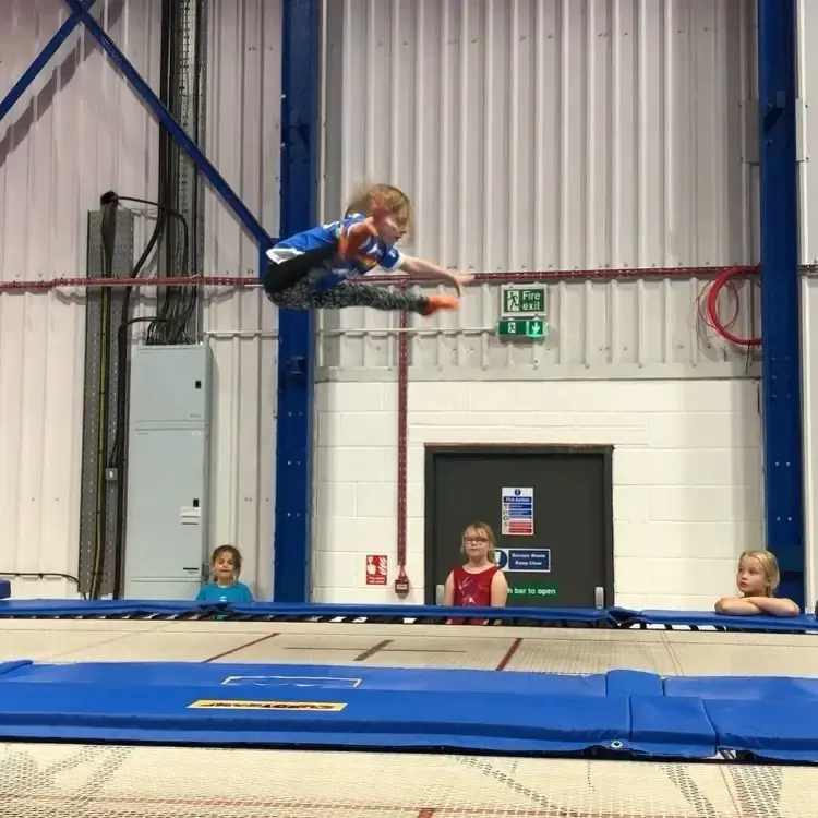 Child performing a horizontal balance move on a rope traverse above a trampoline, with other children watching nearby.