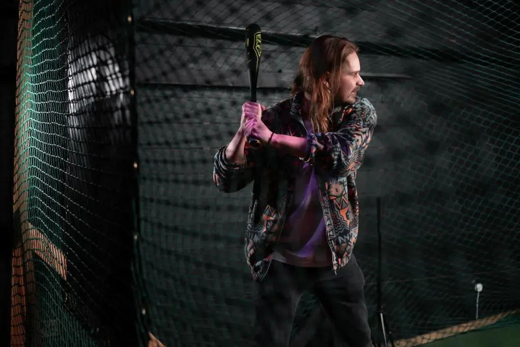 Woman in batting cage holding baseball bat, ready to swing, wearing pink gloves and patterned jacket.