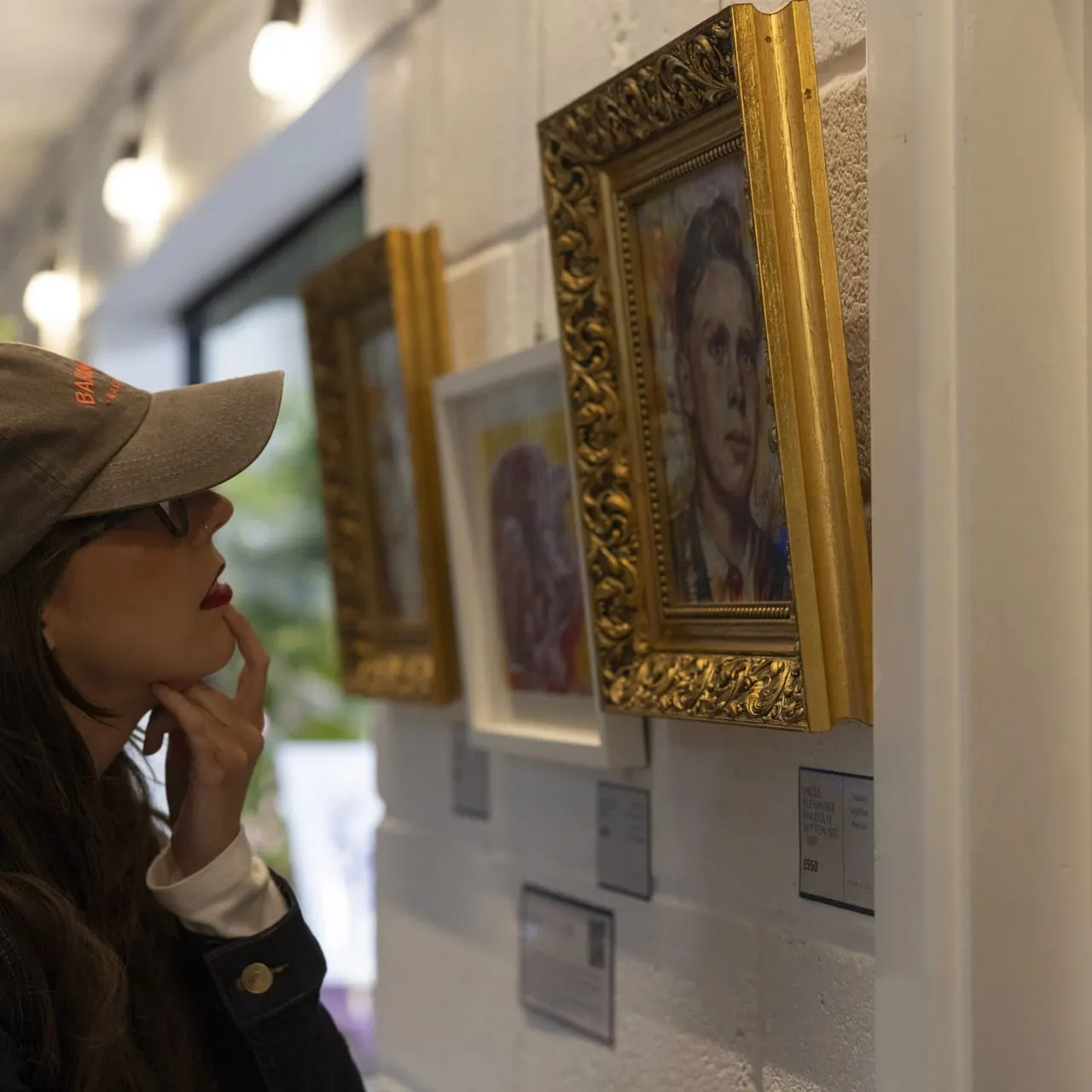 Woman in cap viewing framed portrait paintings in an art gallery interior.