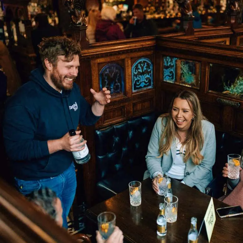 A man and woman laugh together at a bar table with drinks, whilst a staff member serves them in a wood-panelled pub interior.