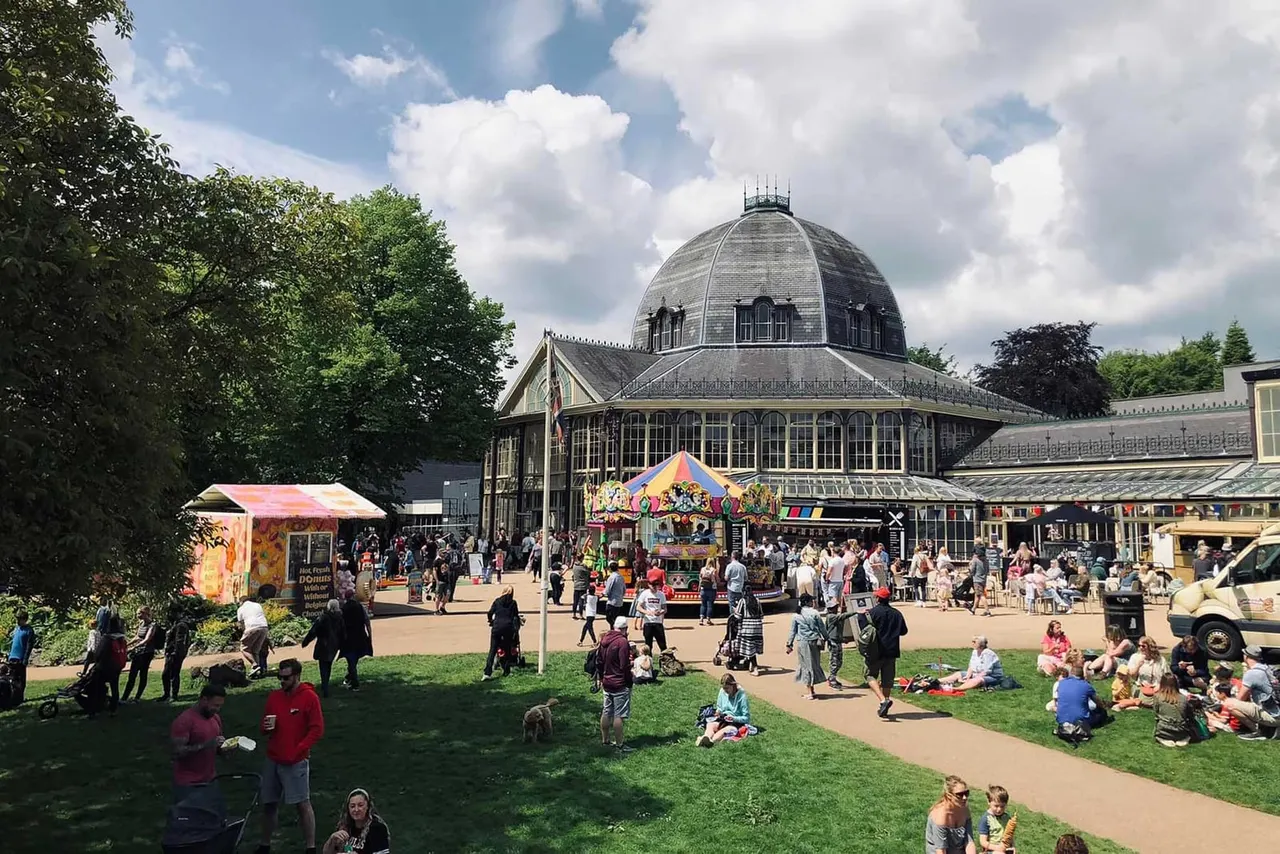 Busy outdoor summer festival with visitors enjoying a fairground carousel and food stalls in front of an ornate domed Victorian building.
