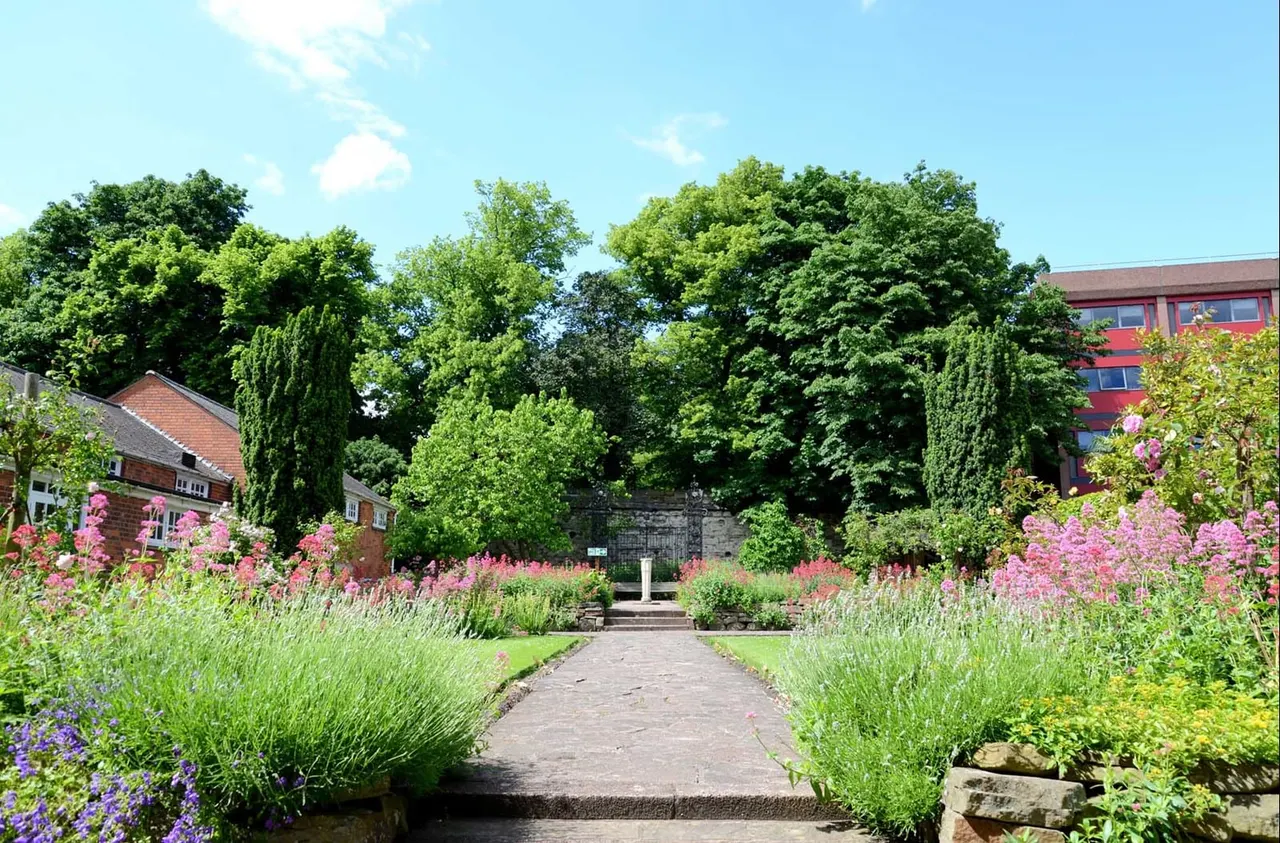 A formal garden path lined with pink flowering plants and lavender leads towards historic brick buildings surrounded by mature trees.