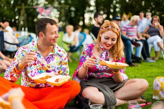 A man and woman in colourful shirts sit on grass eating pizza at an outdoor gathering with spectators in the background.