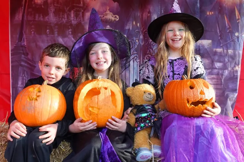 Three children in Halloween costumes holding carved jack-o'-lanterns with a teddy bear against a spooky castle backdrop.