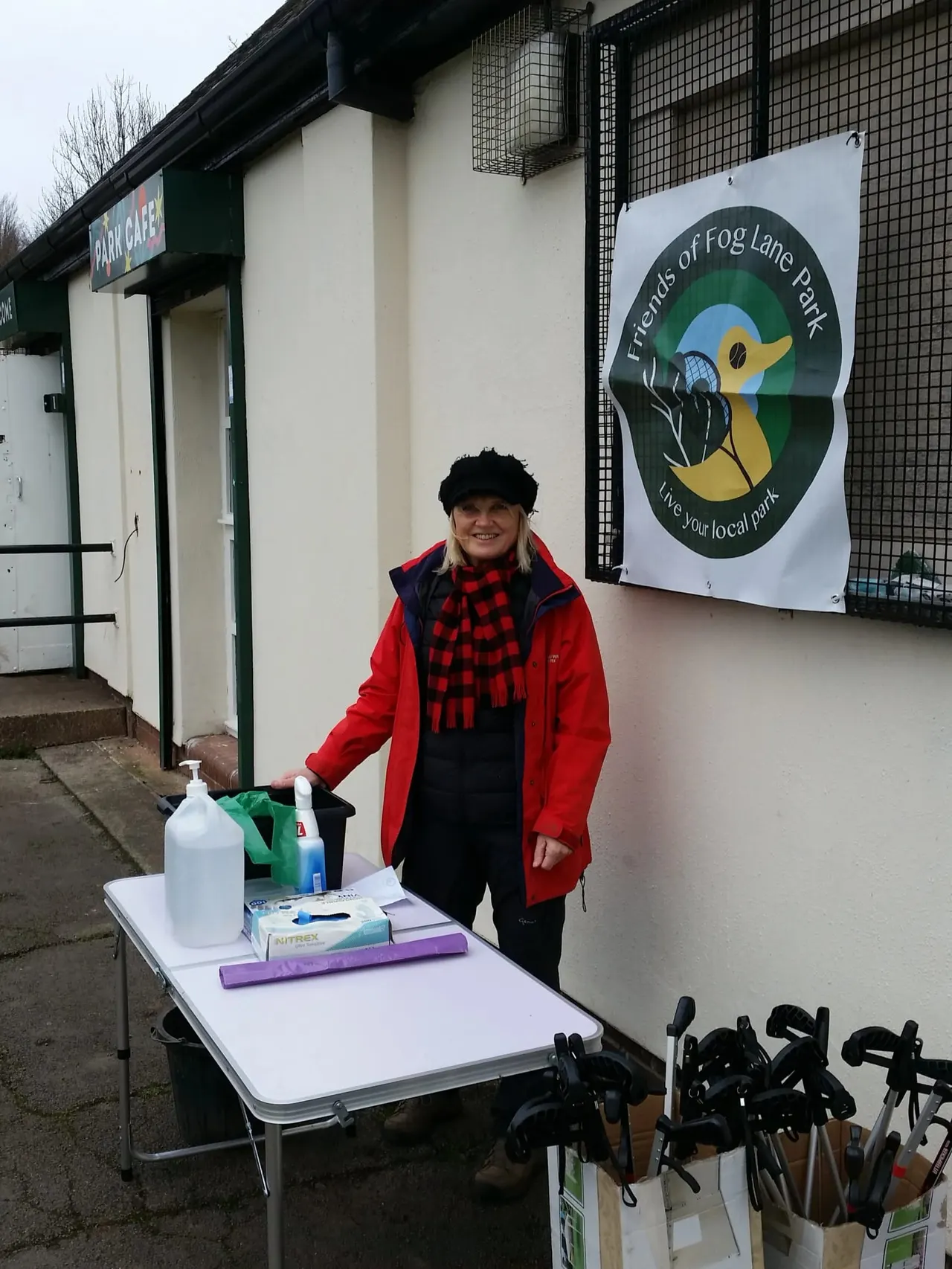 Person in red jacket and black hat standing at an outdoor table with sanitiser and supplies outside a park café.