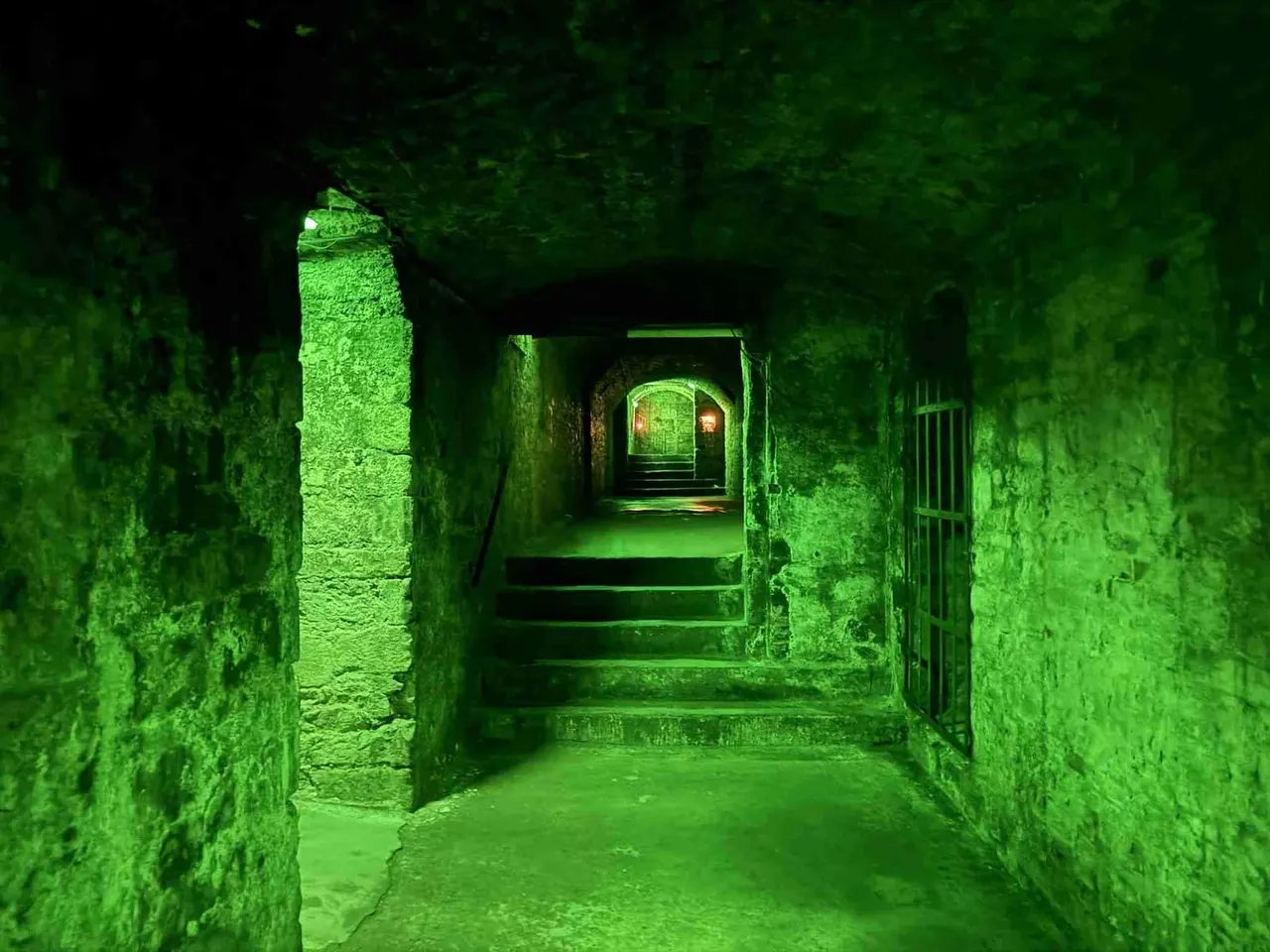 Underground stone passageway lit with green lighting, showing a long corridor with arched doorways receding into the distance.