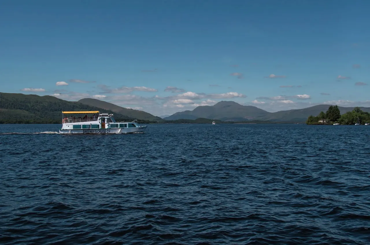 Cruise boat on Loch Lomond with mountains and forested shores in the background under a clear blue sky.