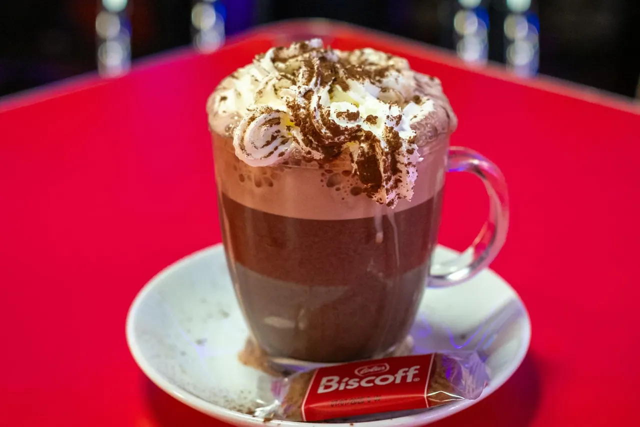 Hot chocolate drink topped with whipped cream and chocolate shavings, served on a red table with a biscuit.