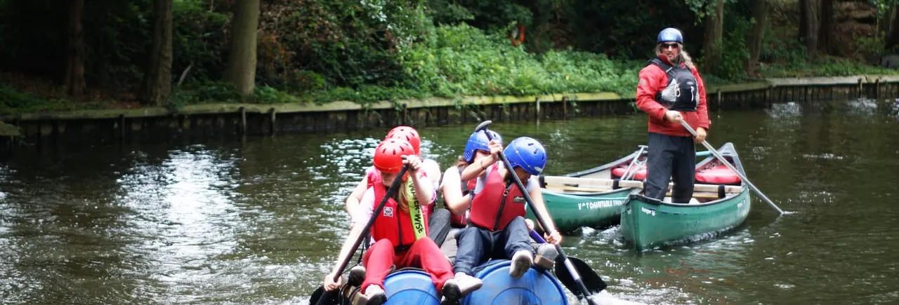 Group of people in safety helmets paddling inflatable boats on a tree-lined river with an instructor in a canoe.