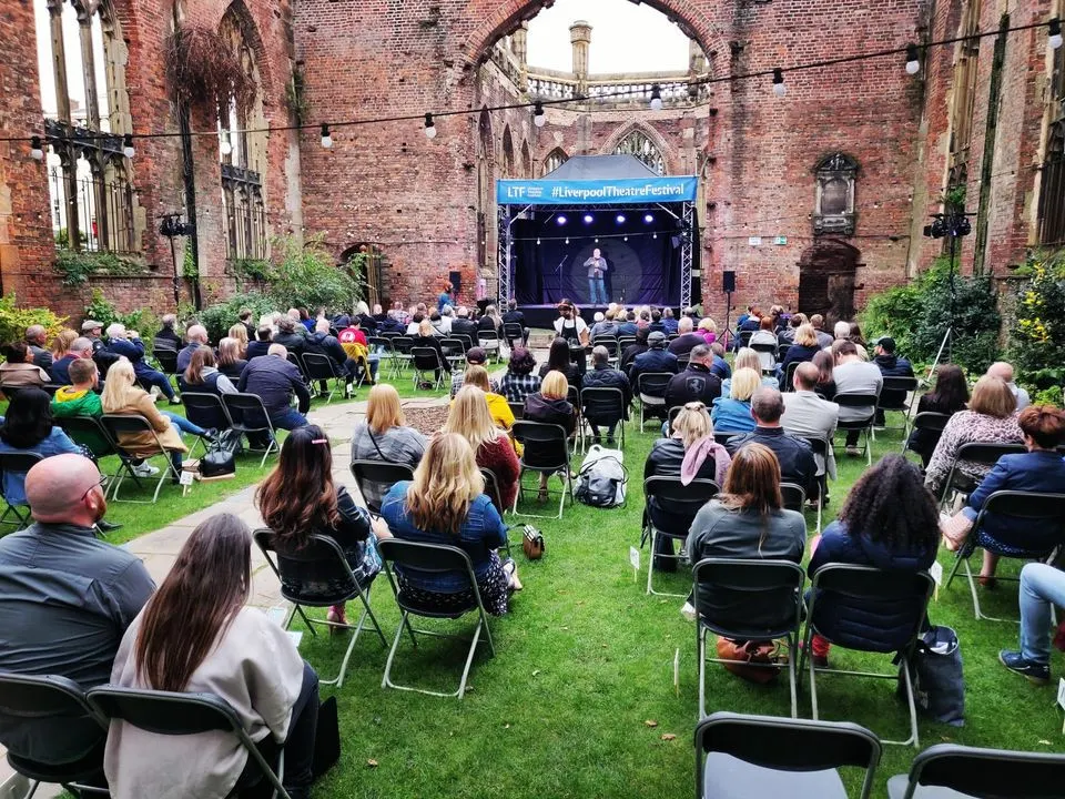 Audience members seated on chairs in a ruined brick church courtyard watching a performance on an illuminated stage.