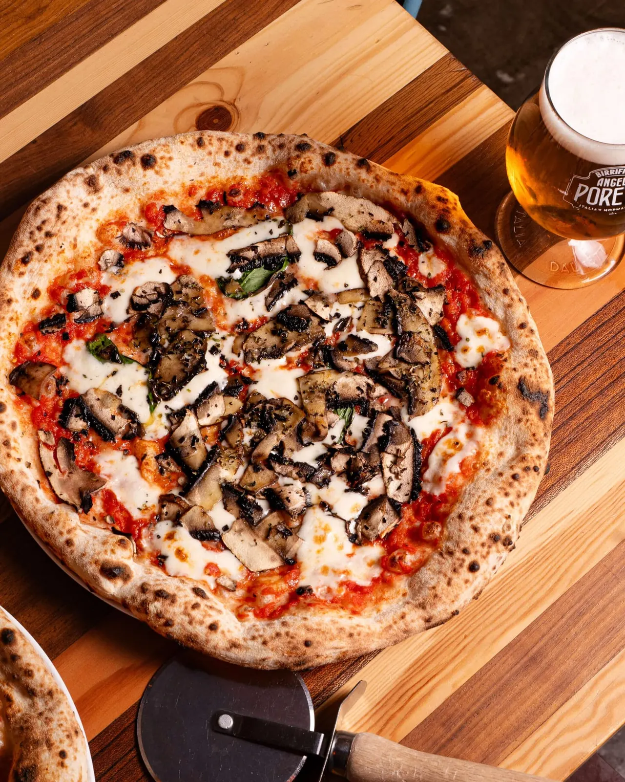 Wood-fired pizza topped with mushrooms, mozzarella and tomato sauce, served with a glass of beer.