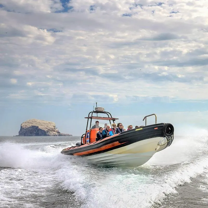 Speedboat full of passengers approaches shore through waves with rocky island visible in background.