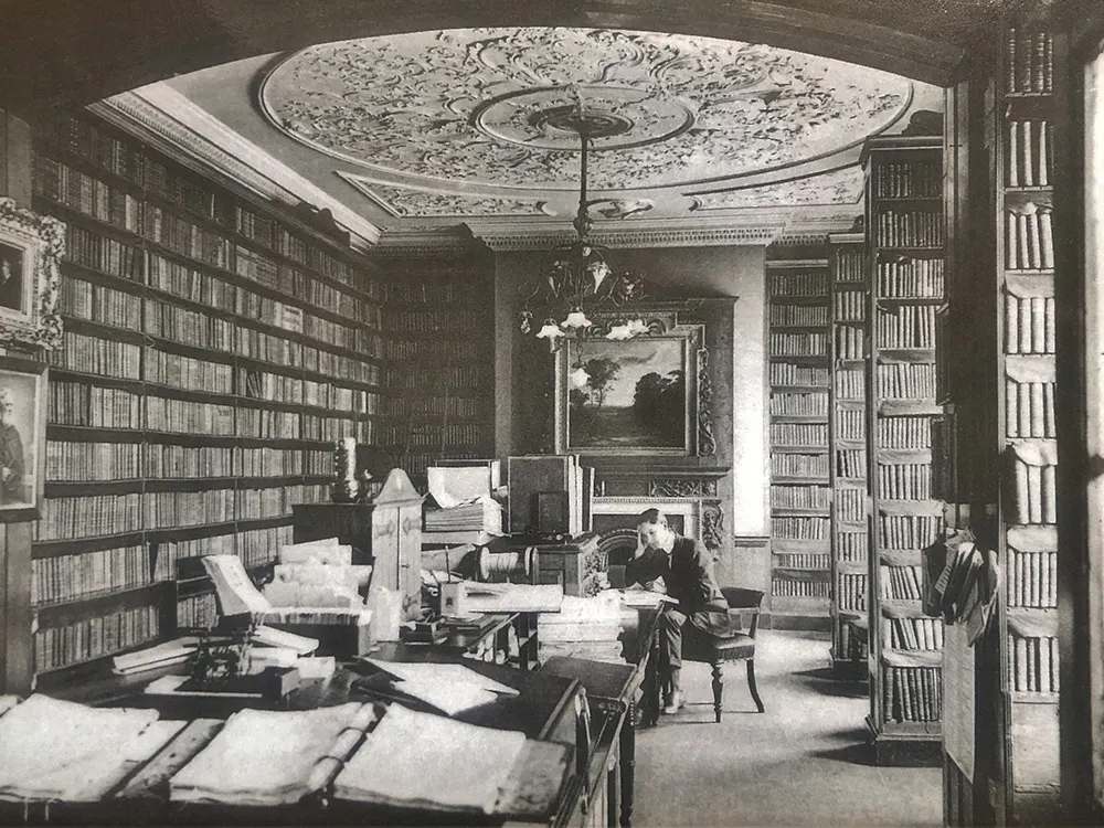 A man sits at a desk in an ornate library room lined with floor-to-ceiling bookshelves and an elaborately decorated ceiling.