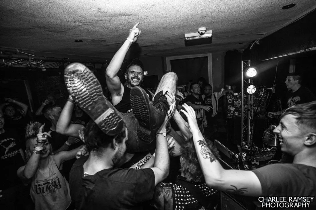 Crowd of people cheering as a man is lifted up during an indoor music event in a basement venue.