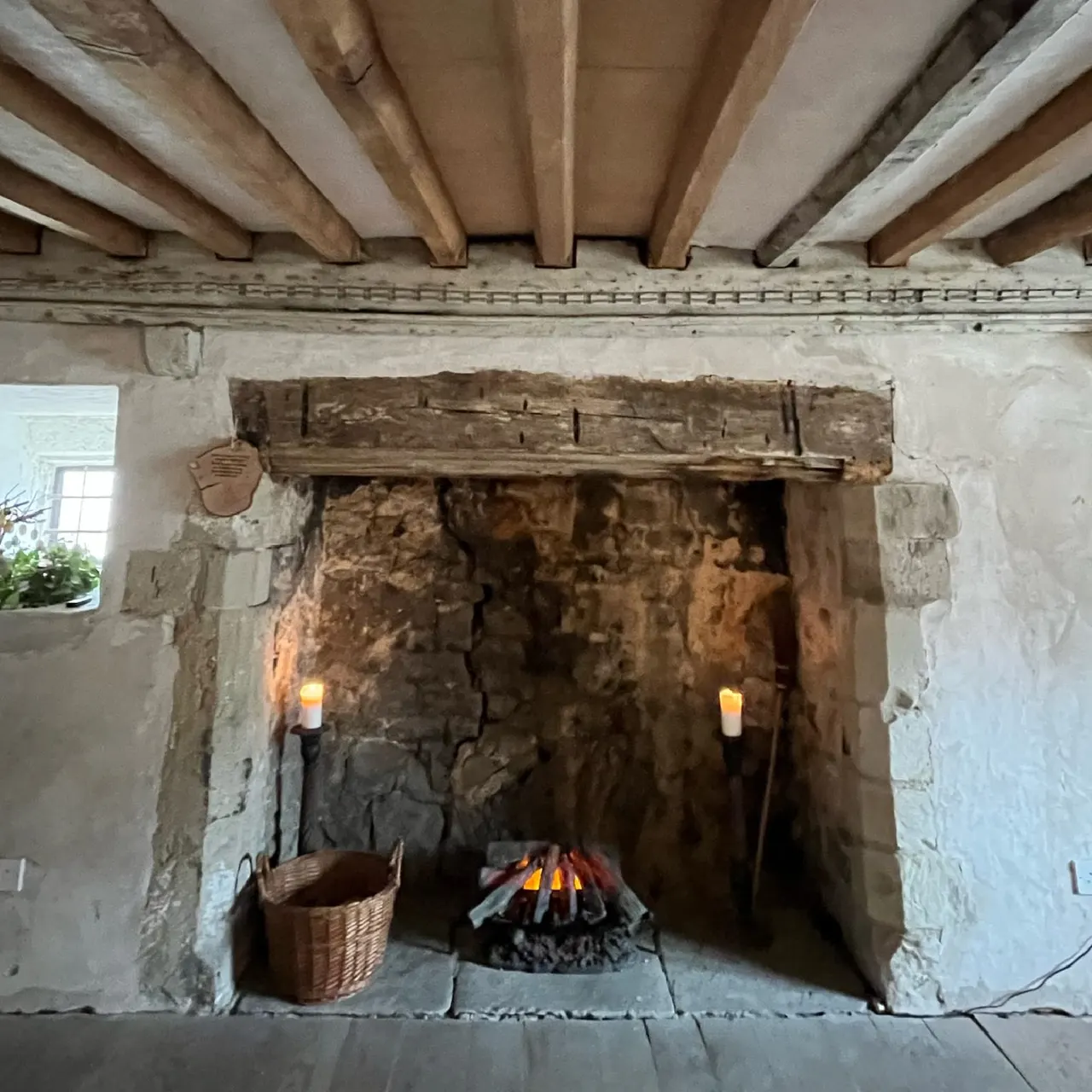 Historic stone fireplace with lit fire, wooden beam ceiling, and wicker basket beside hearth.