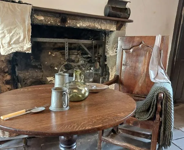 Historic cottage interior with round wooden table, period furniture, and traditional fireplace.