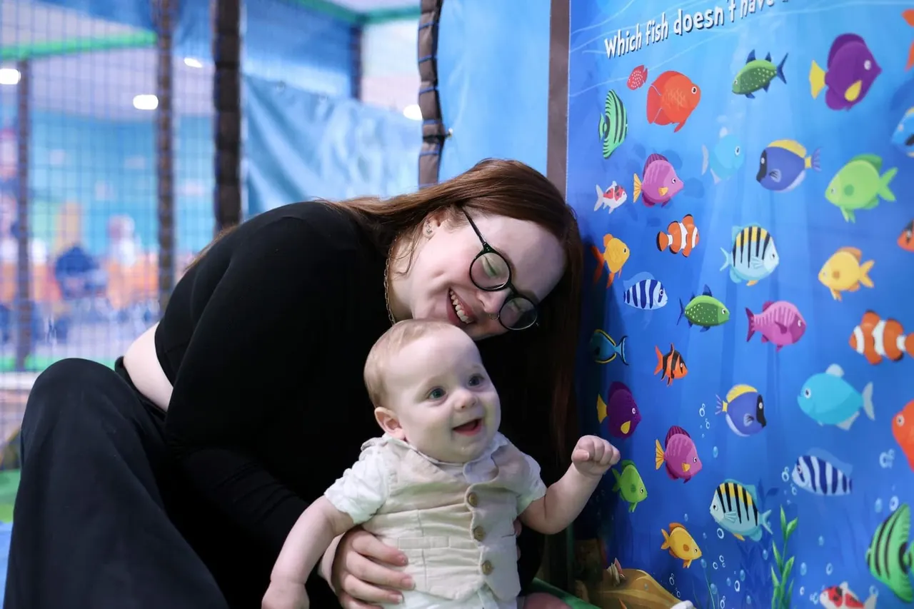 Woman and baby smile in front of a blue interactive panel displaying colourful illustrated fish at an indoor play venue.
