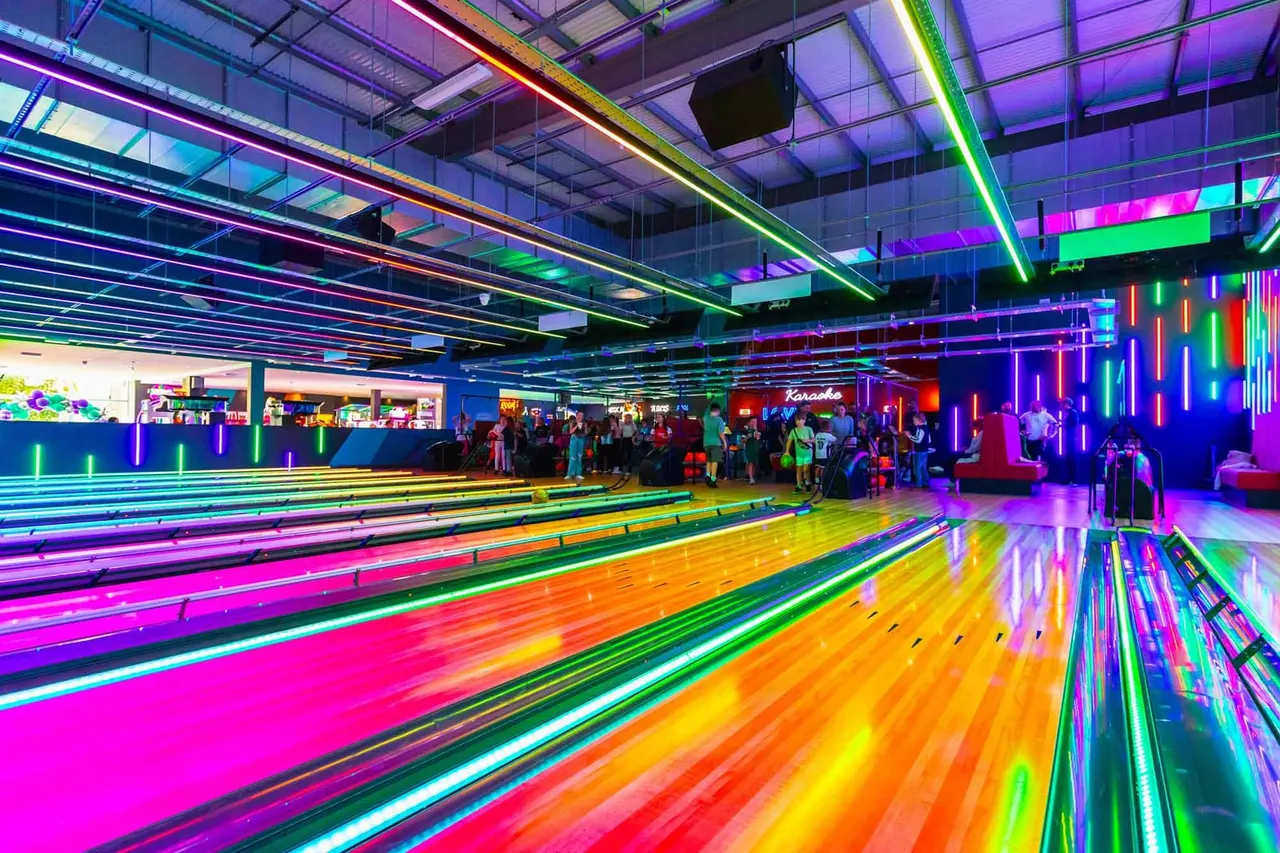 Bowling lanes illuminated with vibrant neon lighting in pink, green, and orange, with visitors standing and socialising in the brightly lit venue.