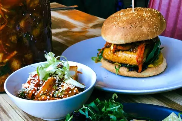 Burger with sesame seed bun and roasted vegetables on blue plate, beside bowl of salad with garnish and soft drink.