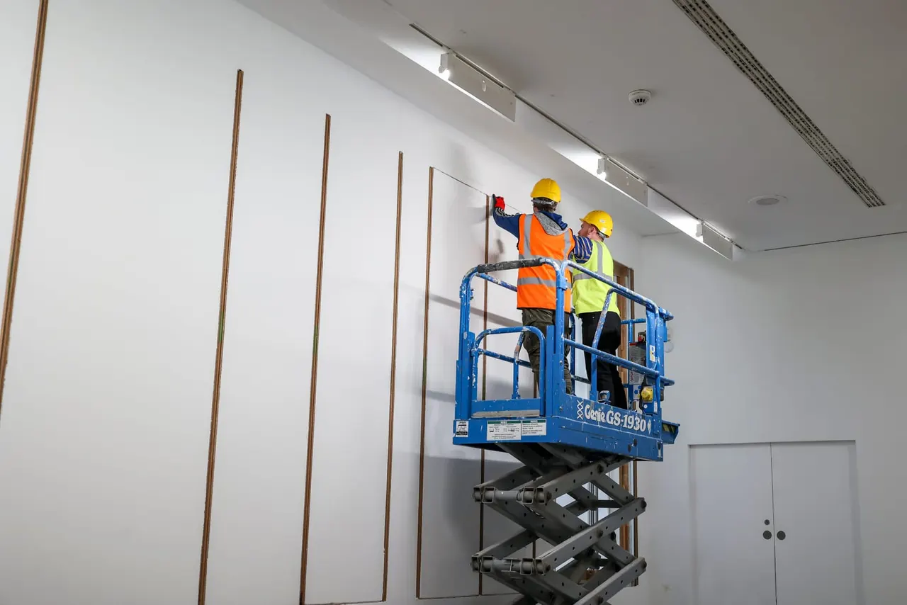 This is a photograph, not a designed graphic.

Two workers in hard hats and high-visibility vests on a blue scissor lift installing or maintaining wall panels indoors.
