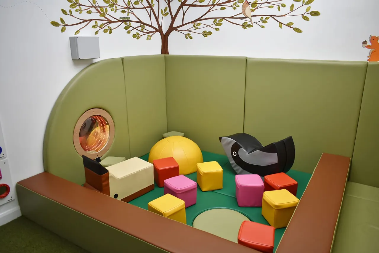 Soft play area with colourful foam blocks, a yellow ball, and play equipment inside a padded green enclosure decorated with a tree mural.