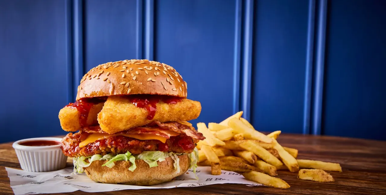 A gourmet burger with fried chicken, bacon and cheese on a seeded bun, served with chips and sauce.