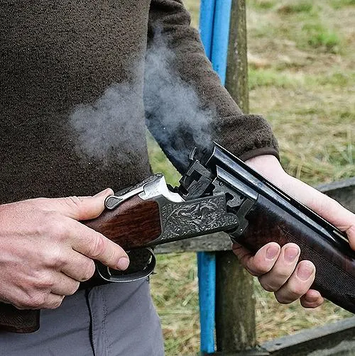 Person firing a shotgun at an outdoor shooting range, with smoke visible from the discharge.