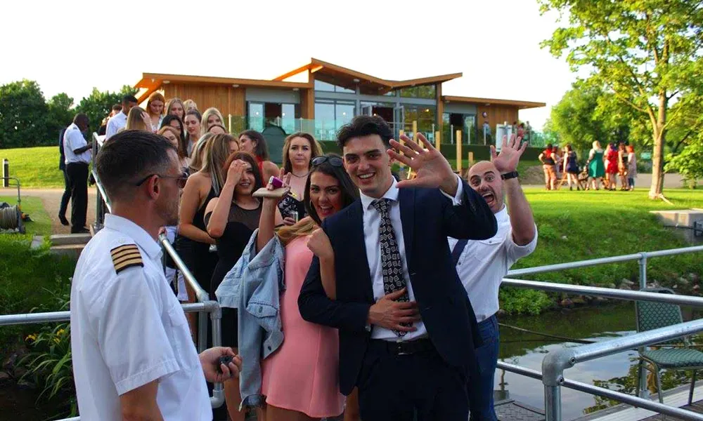 A group of smartly dressed guests enjoying a celebration on a riverbank, with a modern wooden pavilion building visible behind them.