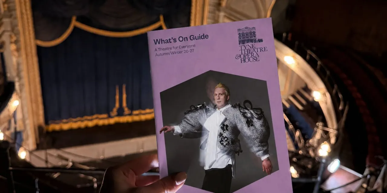 A hand holds a lavender programme for the Tyne Theatre & Opera House in front of an illuminated theatre mirror.