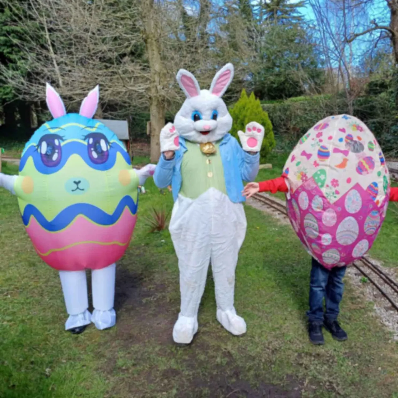 Person in Easter bunny costume posing with two people in colourful Easter egg costumes outdoors on a path with trees.